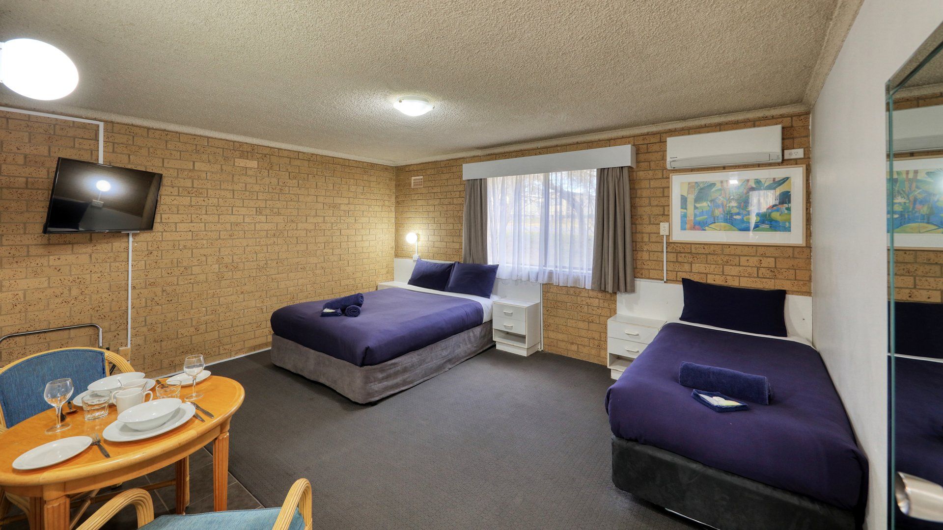 Small hotel room with two beds, dining table, TV, and window with curtains.