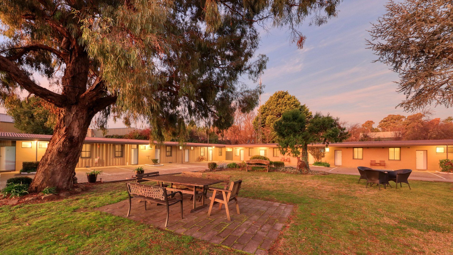 Courtyard of single-story buildings at sunset with picnic tables, a large tree, and warm lights