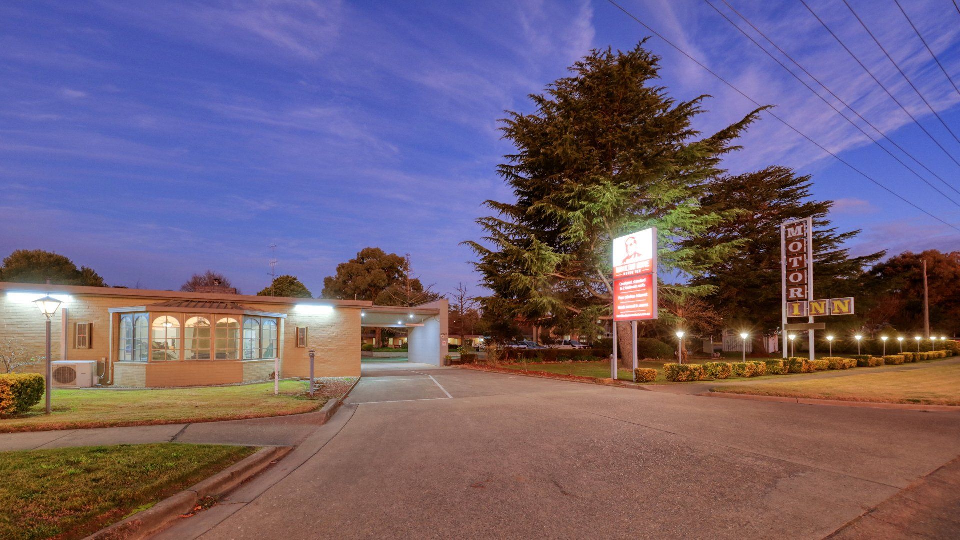 Dusk view of a motel and parking lot with lit signs, trees, and a blue-purple sky