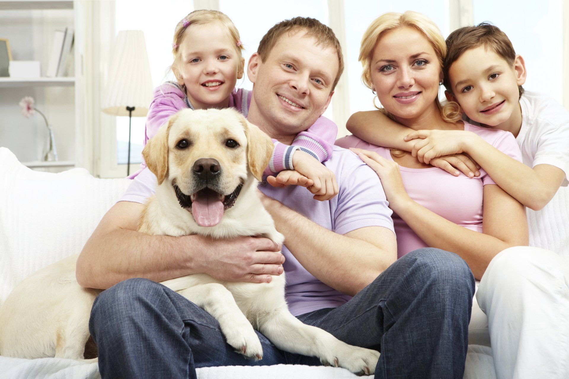 Family, including a dog, smiles on a couch. The dog is held by the man, the woman and children hug.