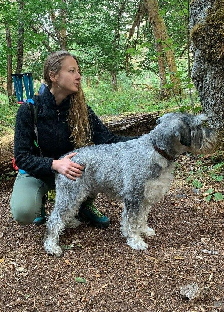 Woman kneeling beside a gray and white dog in a forest, petting its back.