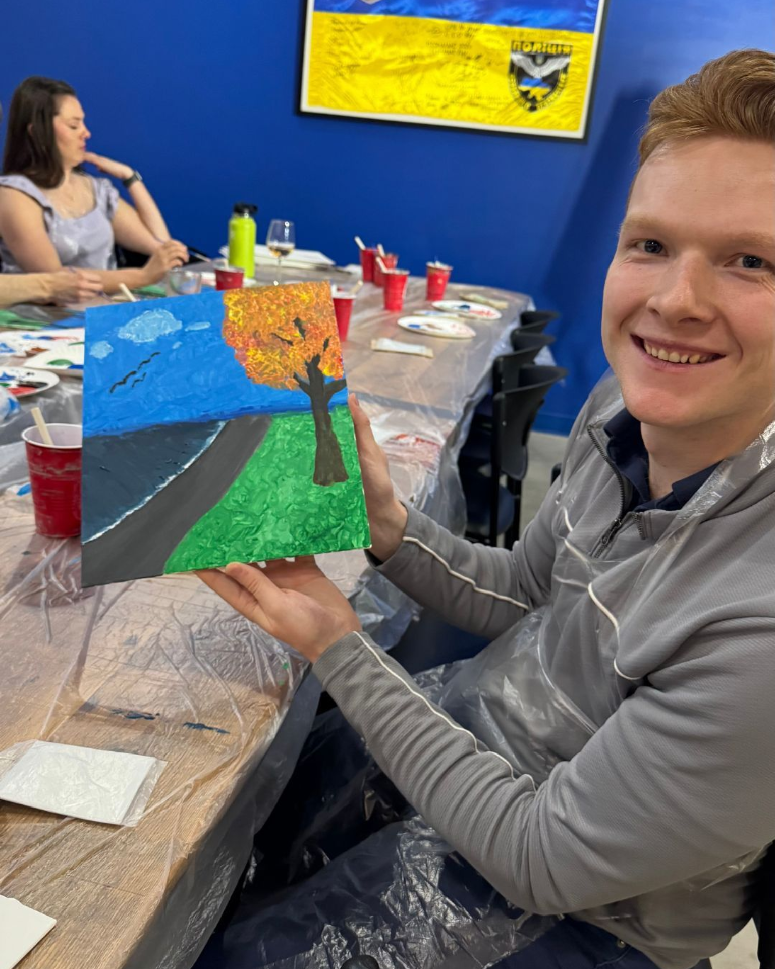 Person holding a colorful painting at a table during an art activity, smiling in a bright room.
