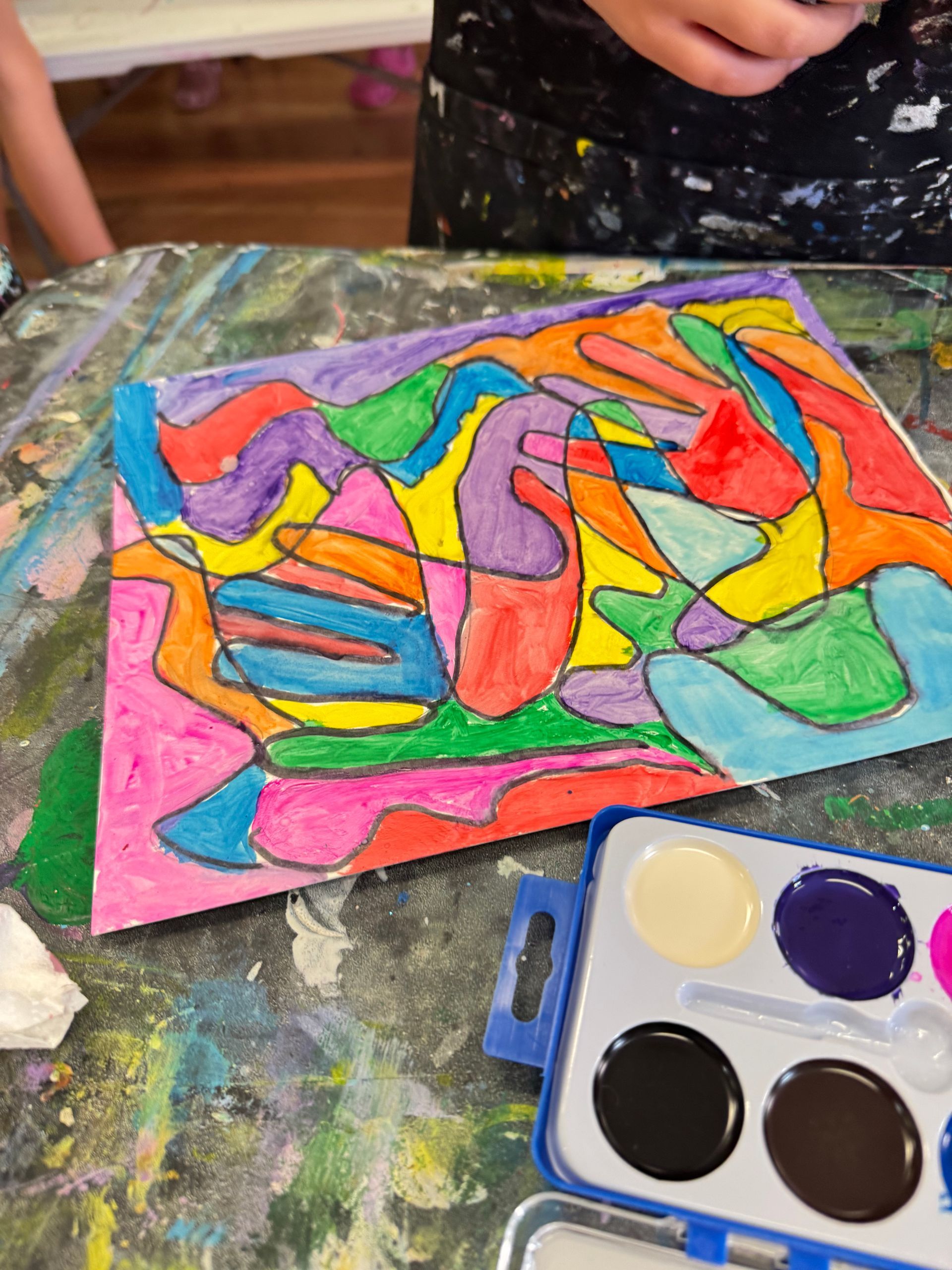 Colorful abstract painting on paper beside a watercolor palette, with a child’s hands nearby