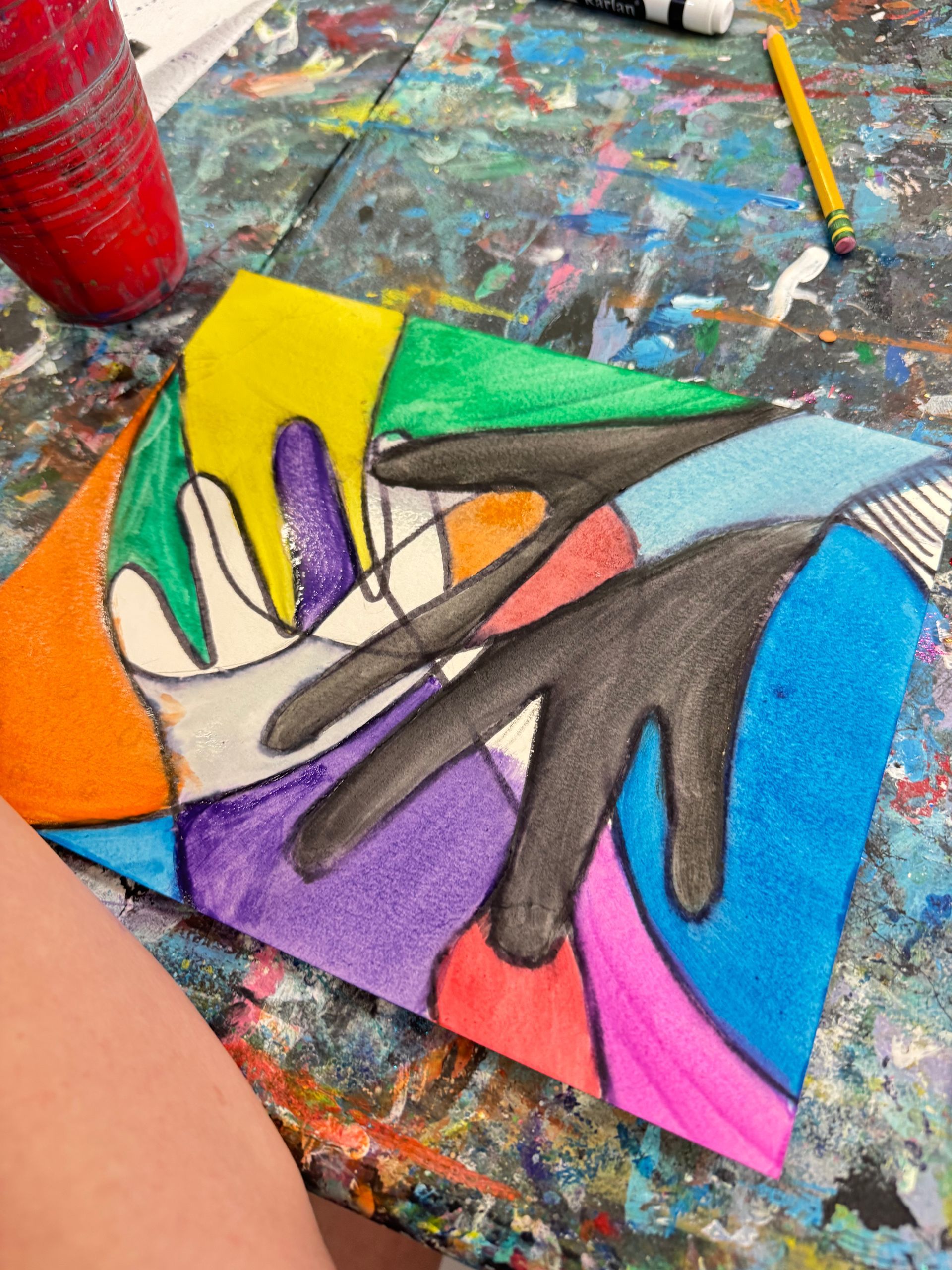 Colorful abstract painted hands artwork on a small canvas, surrounded by art supplies on a paint-splattered table