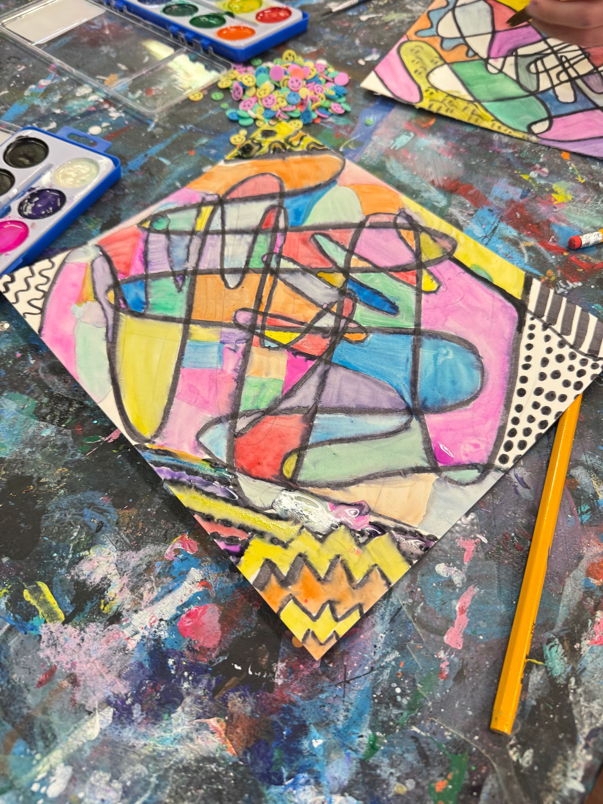 Colorful abstract paper collage on a paint-splattered table, with crayons and art supplies nearby