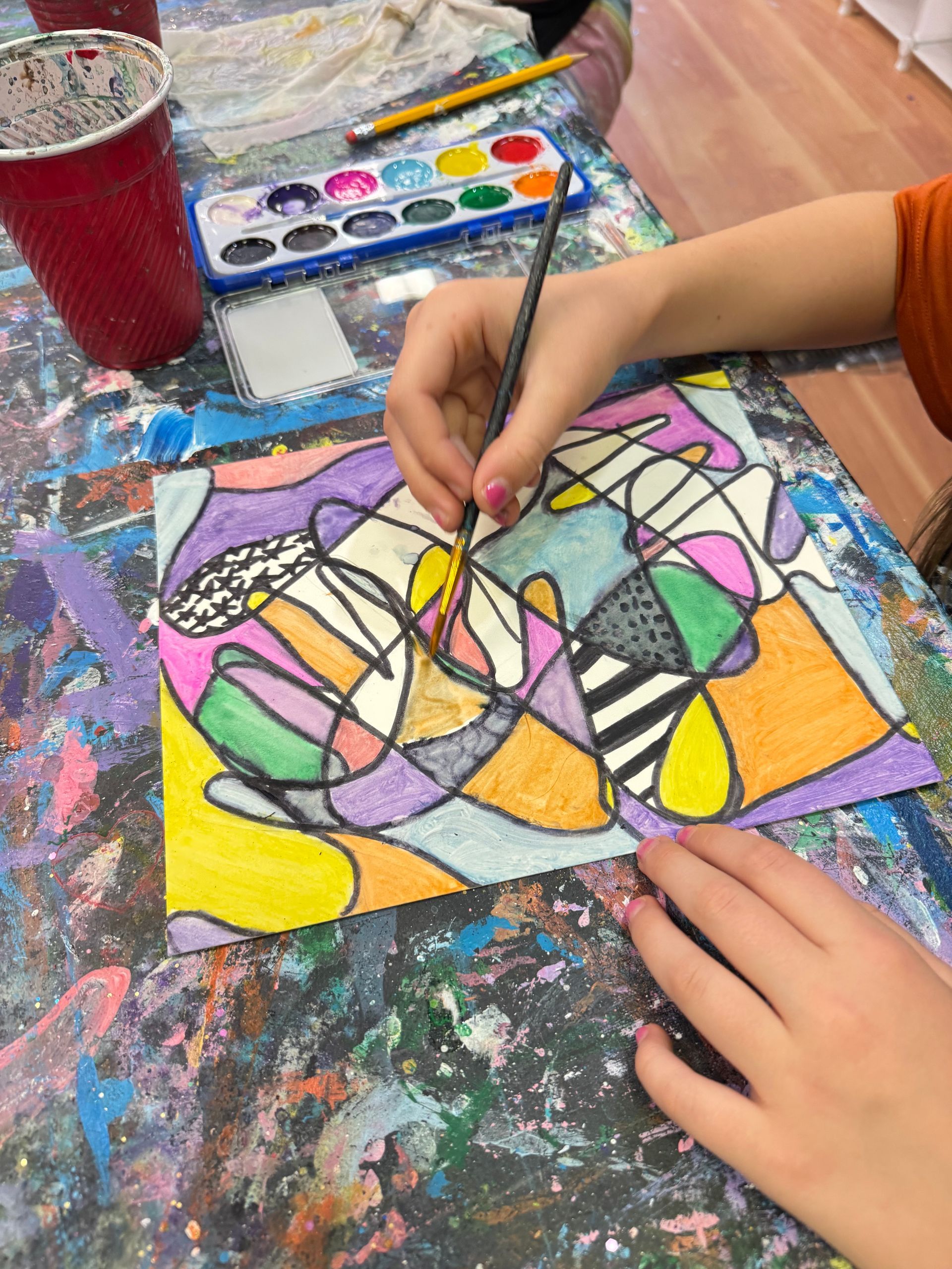 Child painting a colorful abstract design with watercolor on paper at a paint-splattered table