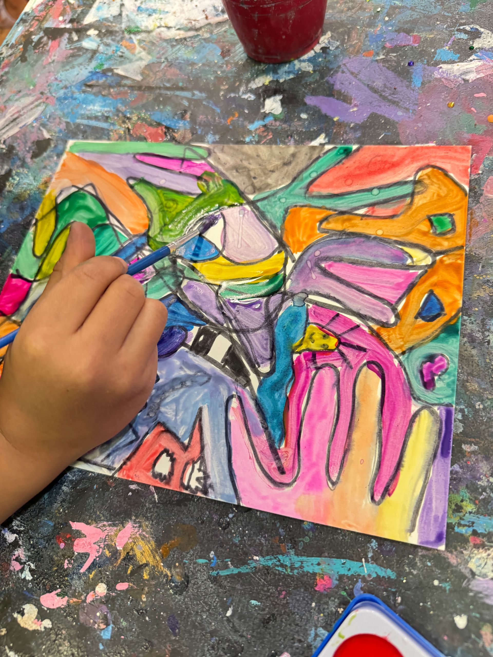 Child painting a colorful abstract artwork with a brush on a paint-splattered table