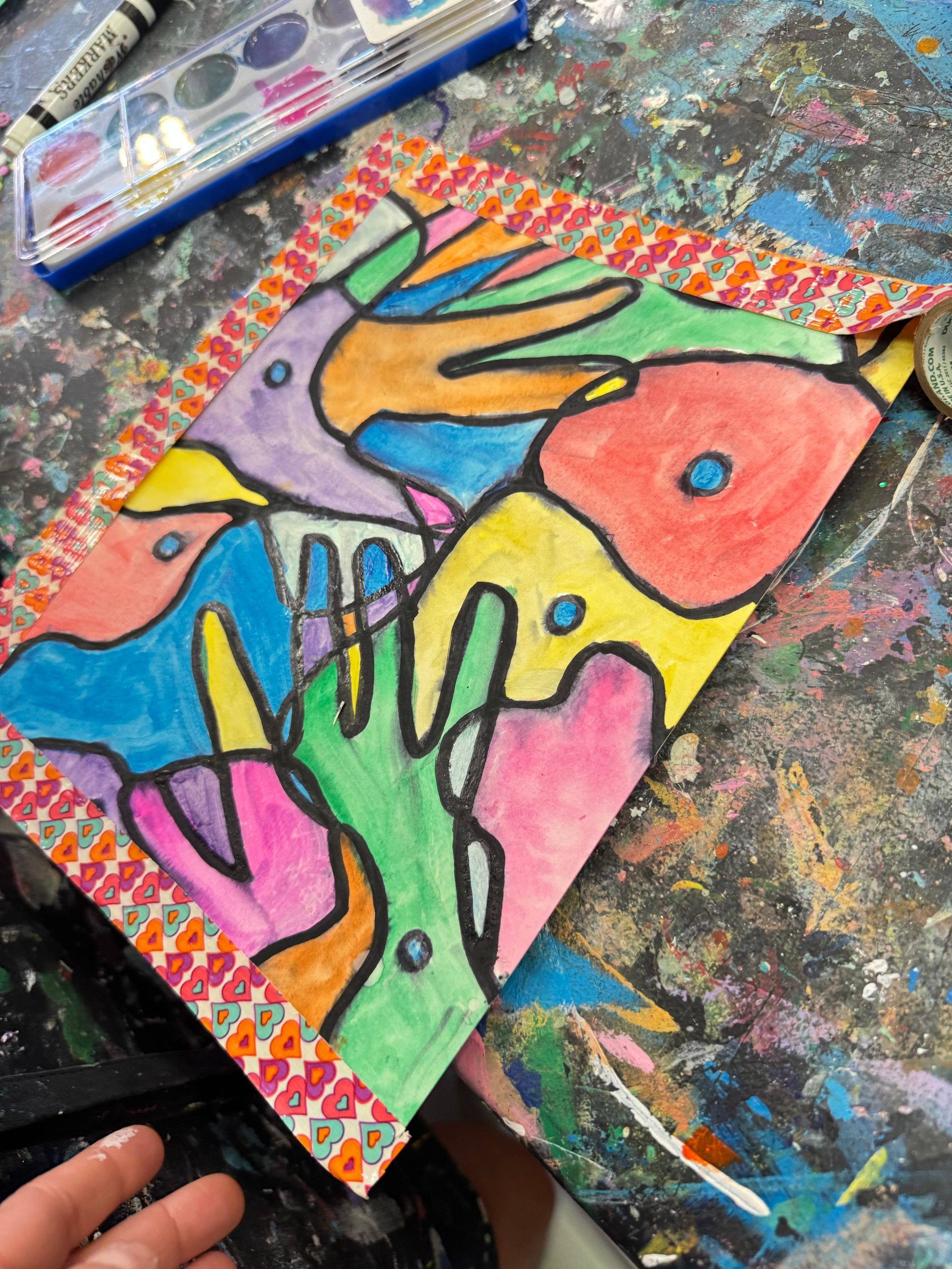 Colorful abstract handprint artwork on paper, with paint supplies and a paint-splattered surface nearby.
