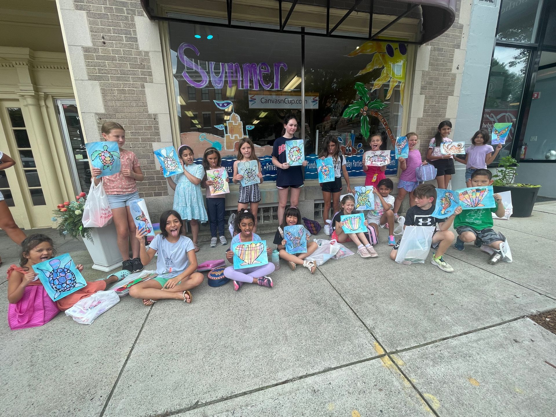 A group of children are sitting on the sidewalk holding paintings.