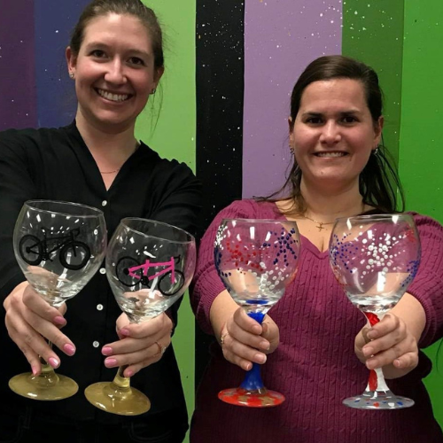 Two women holding wine glasses they decorated, in front of a colorful backdrop.
