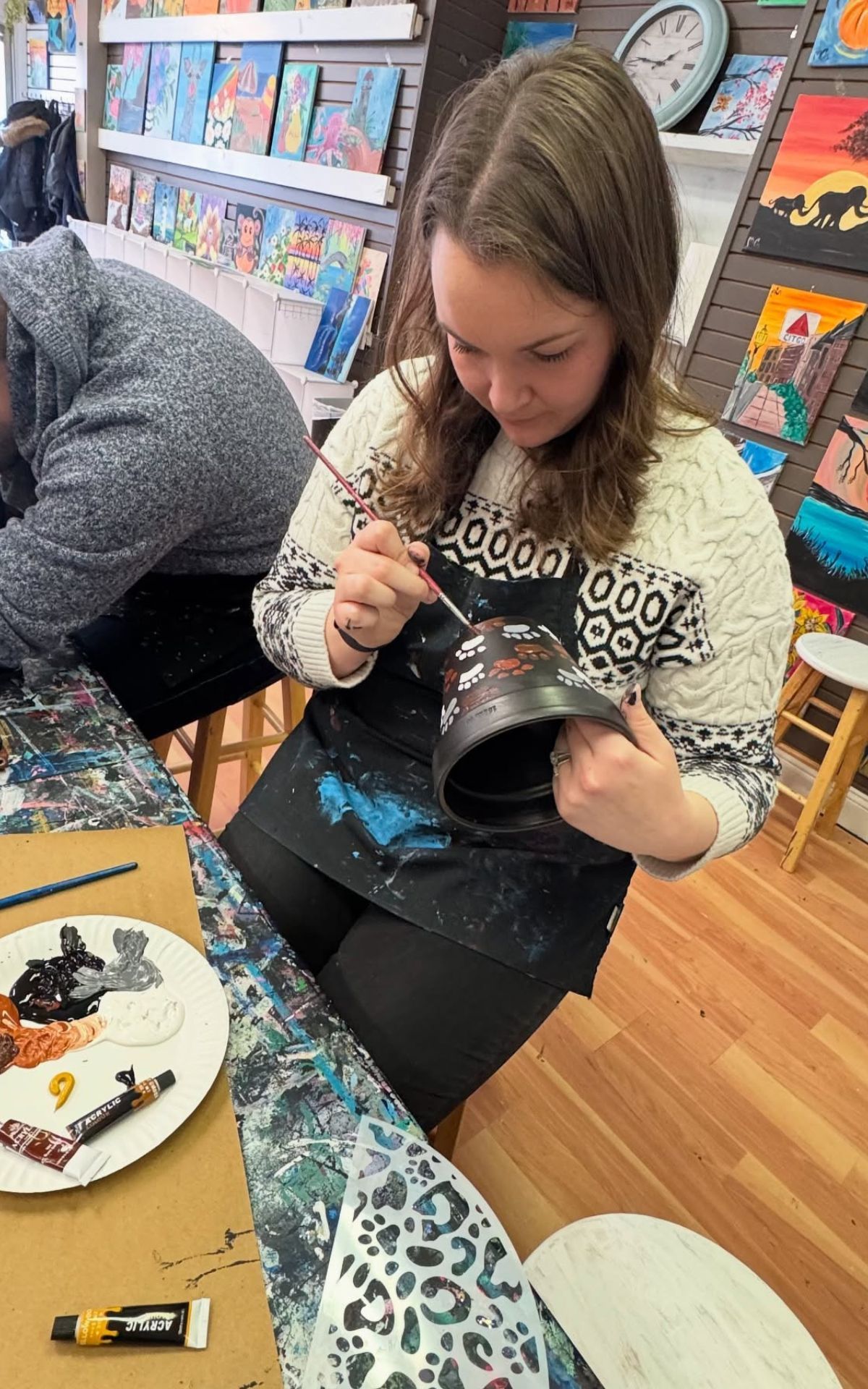 Woman painting a ceramic pot in a studio. She wears a sweater and apron, using a paintbrush.