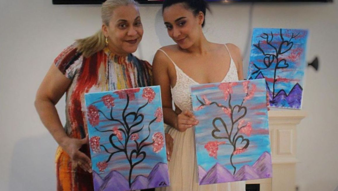 Two women holding paintings of trees with heart-shaped leaves, smiling. Paintings feature pink, purple, and blue colors.