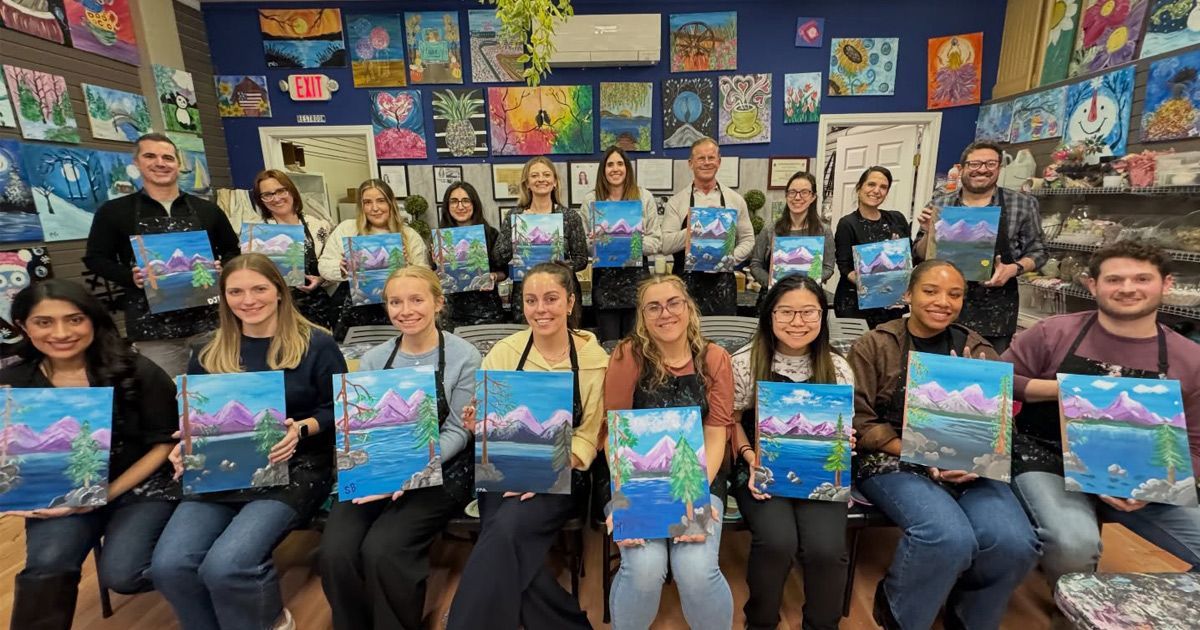 Group of people seated in a classroom, holding colorful abstract paintings in front of a wall of artwork.