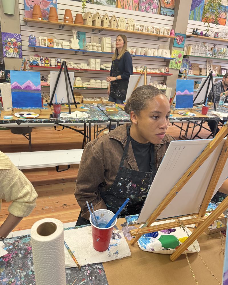 Woman painting at an easel in an art class, with supplies on the table and shelves of artworks behind her.
