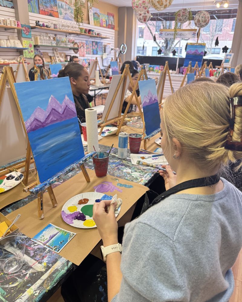 People painting at easels in an art studio, with colorful canvases and supplies on tables.