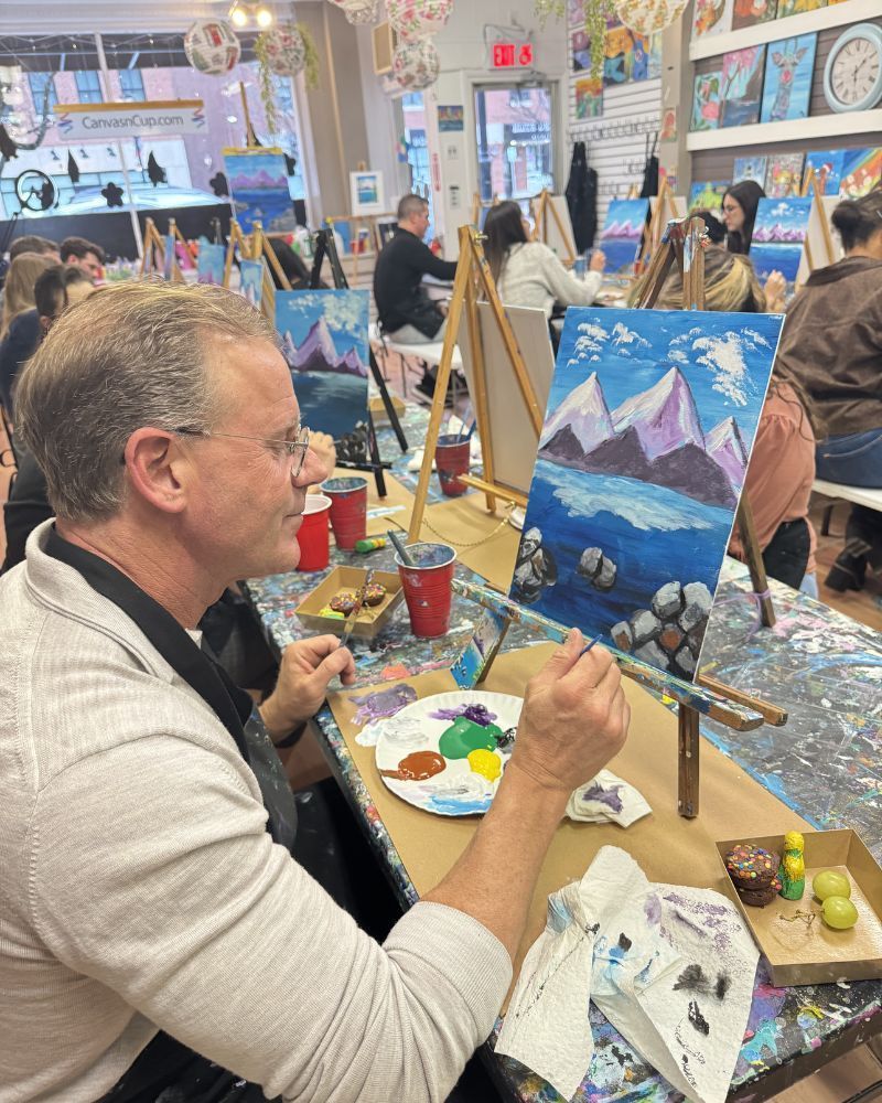 Man painting a snowy mountain scene at an indoor art workshop, with easels, paints, and other participants nearby.