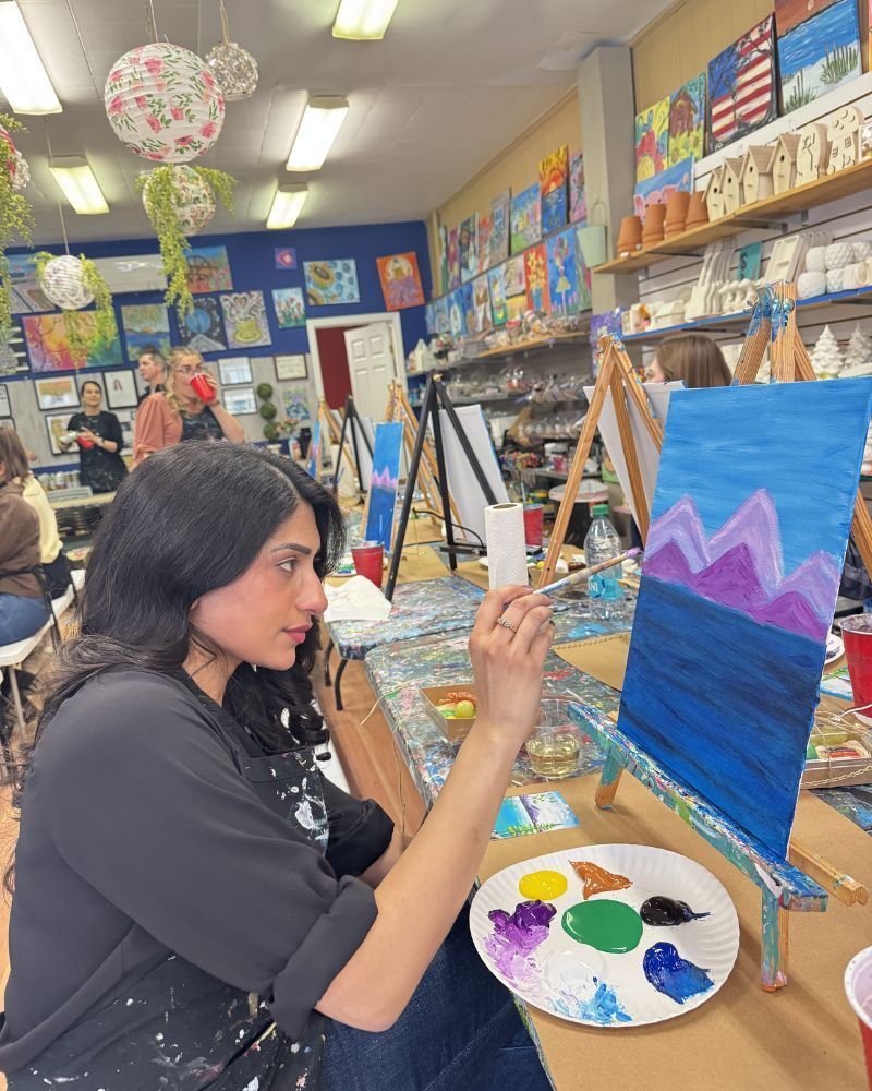 Art class with a person painting a blue mountain scene on an easel, palette on a table.