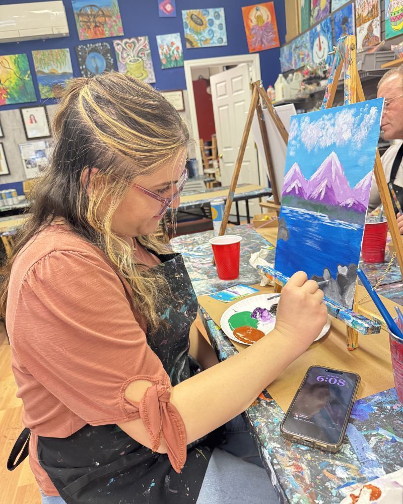 Person painting a mountain landscape on a small canvas in an art studio, with paints and brushes nearby