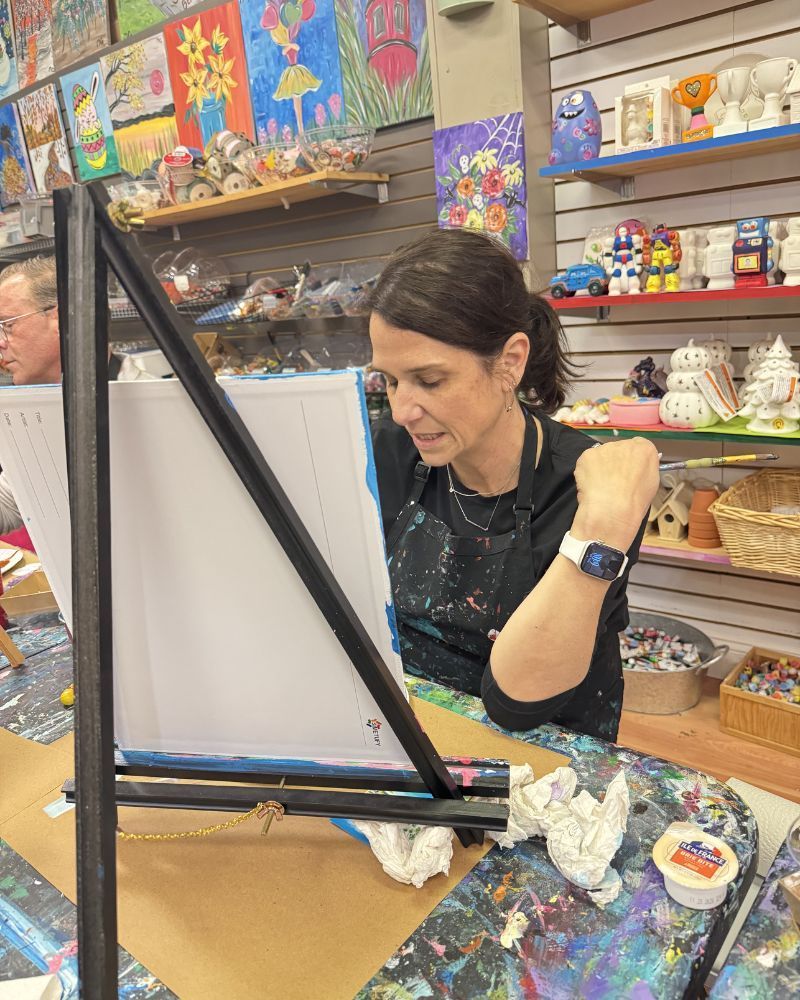 Woman painting at an easel in a colorful art studio, with shelves of crafts behind her.