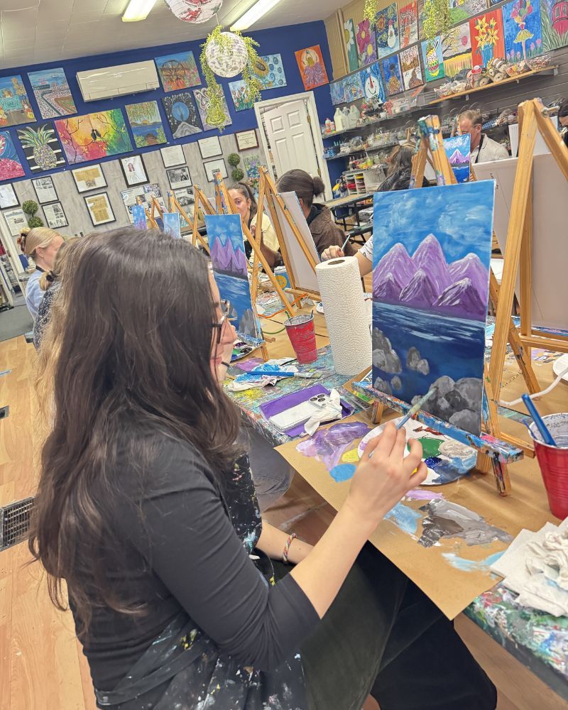 Art student painting a blue mountain landscape at an easel in a colorful studio.