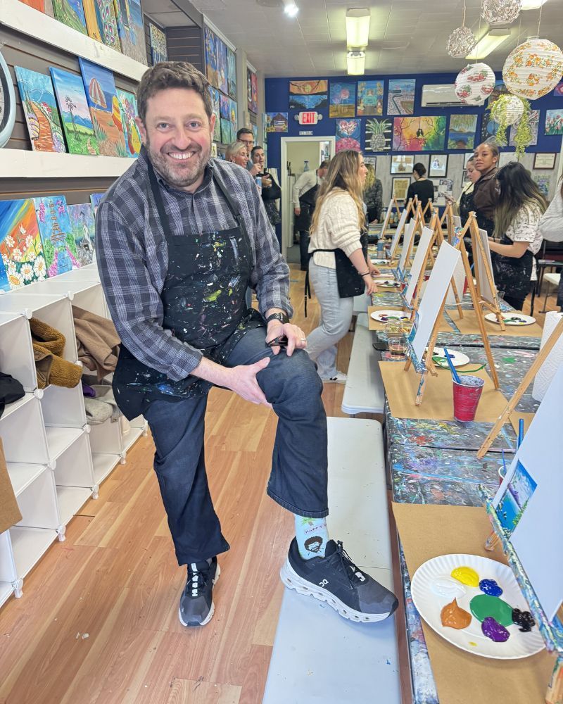 Man smiling in art studio, adjusting his shoe beside colorful paintings and easels.
