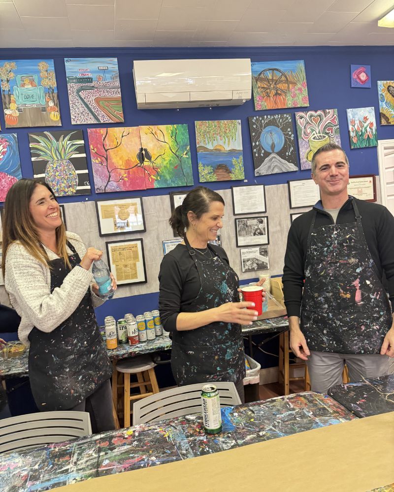 Three people standing behind an art-covered table in a classroom, smiling and talking.