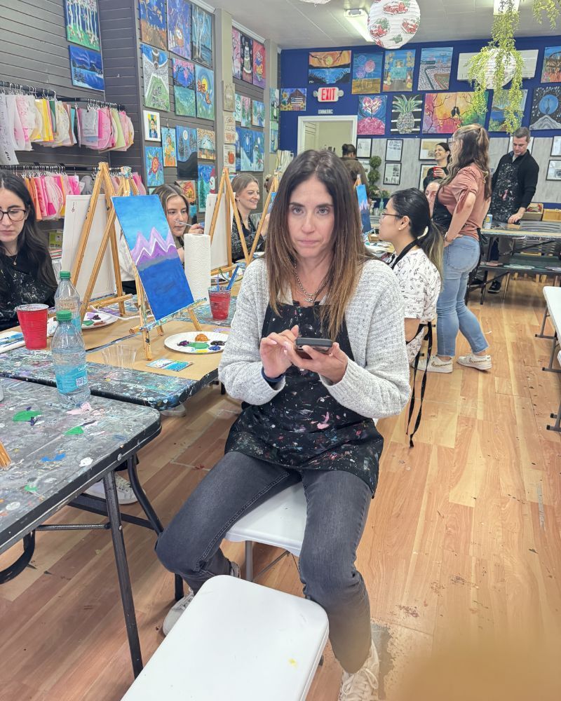 Person seated at an art classroom table, holding a phone, surrounded by paint supplies and other students