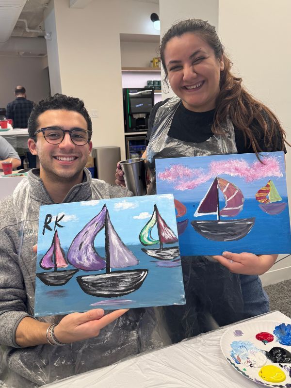 Two people smiling, holding sailboat paintings in an art studio.