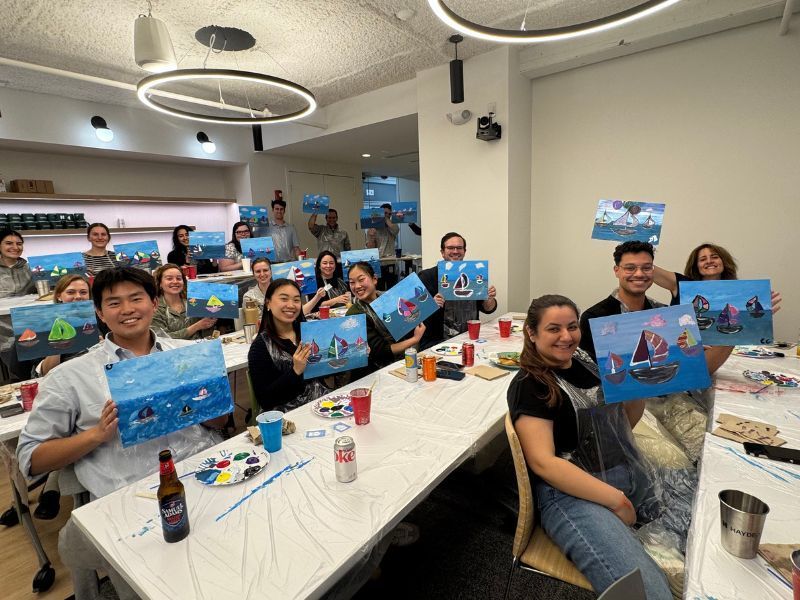 Group of people at a painting party; holding up paintings with blue water and colorful scenery.