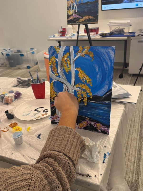 Person painting birch trees with white and yellow swirls at a craft table.