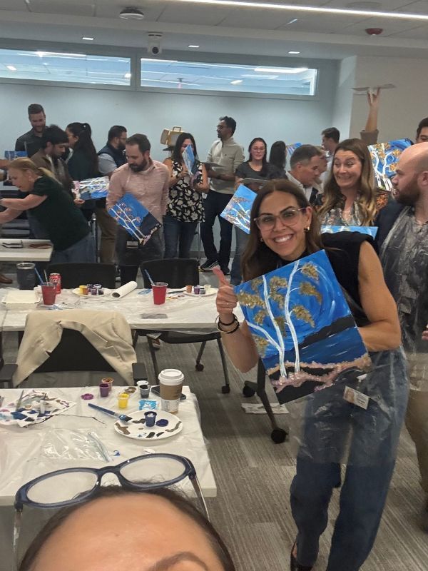 Boston Scientific employees holding painted canvases at a lively indoor art event, smiling and posing among tables and supplies.