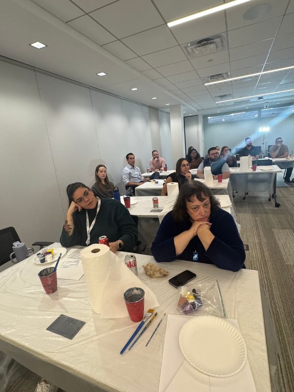  Boston Scientific employees  seated at tables in a bright room, listening with thoughtful expressions watching Mahsa explaining painting techniques.