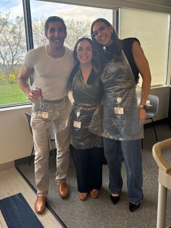 Three  Boston Scientific employees in paint party gear pose smiling in an office with a large window.
