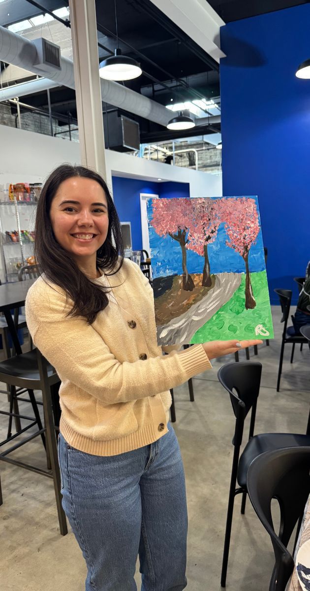 Woman smiles, holding a colorful painting of trees, road, and sky. Inside a building, wearing light sweater and jeans.