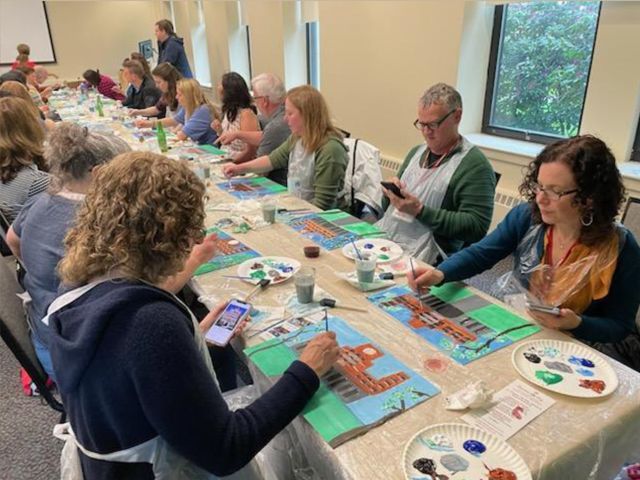 People painting at a long table in a bright room, using colorful art supplies and checking phones.