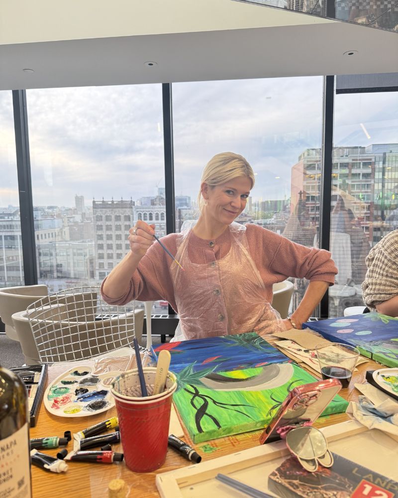 Person painting at a table with colorful artwork and supplies, seated by large windows overlooking a city skyline.