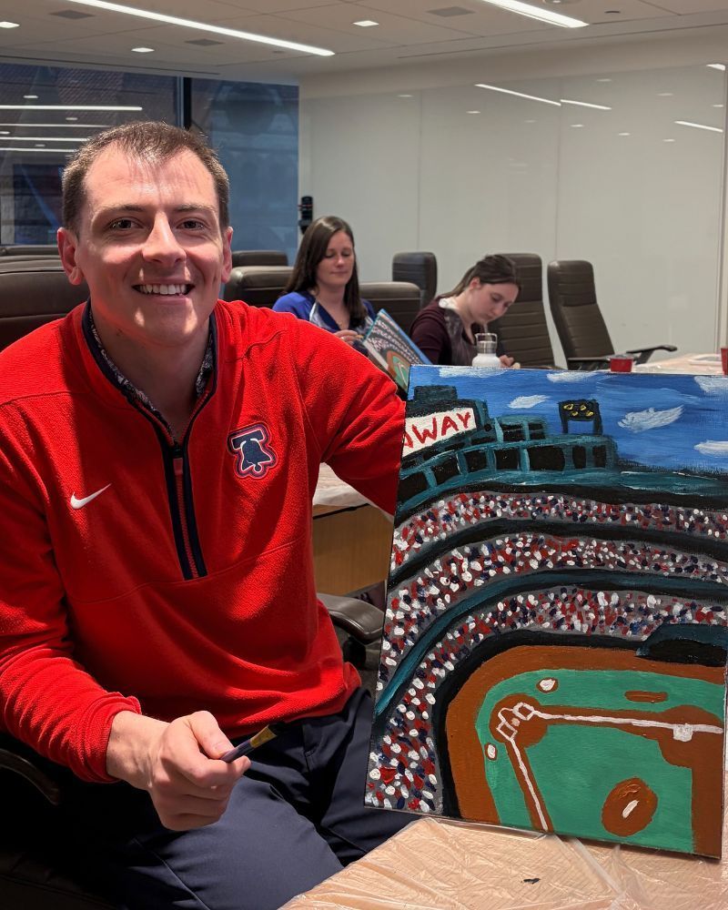 Man in red sweatshirt sitting in an office beside a colorful baseball stadium painting
