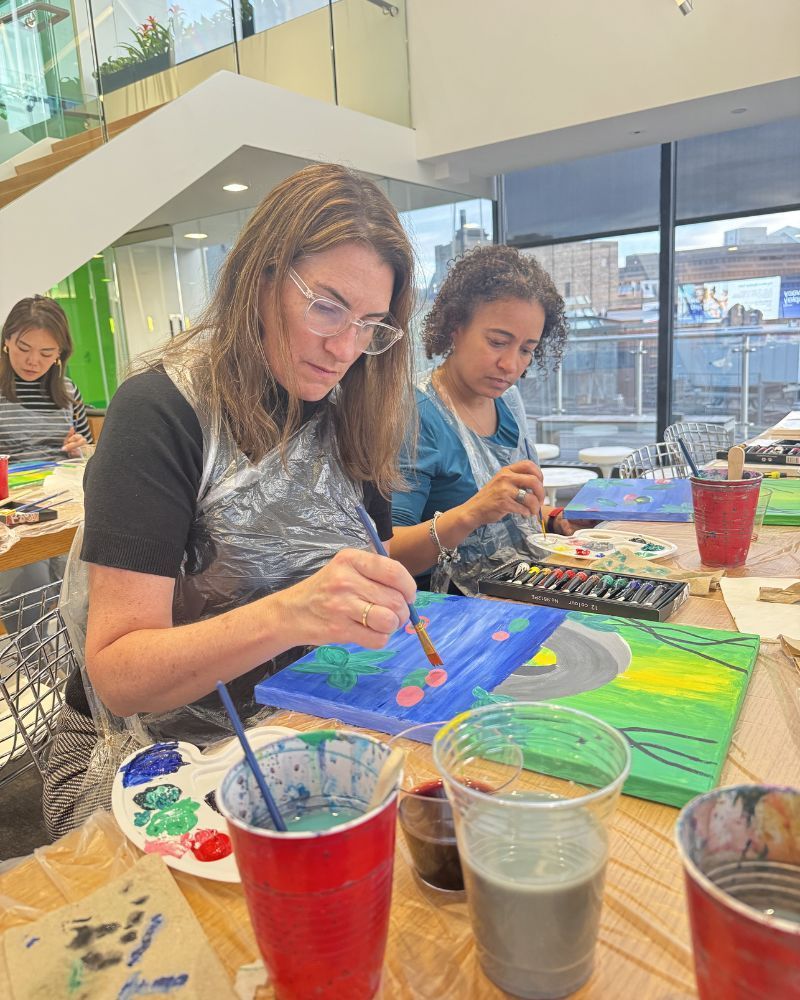 People painting colorful canvases at a table in a bright indoor studio