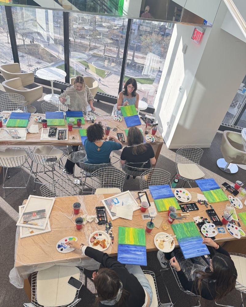 People painting at tables in a bright art studio with large windows and craft supplies.