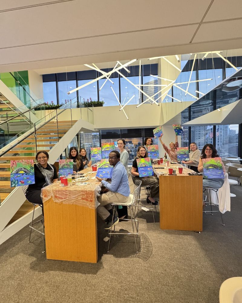 People seated at tables in a bright lobby with books and crafts, near a staircase and large windows.