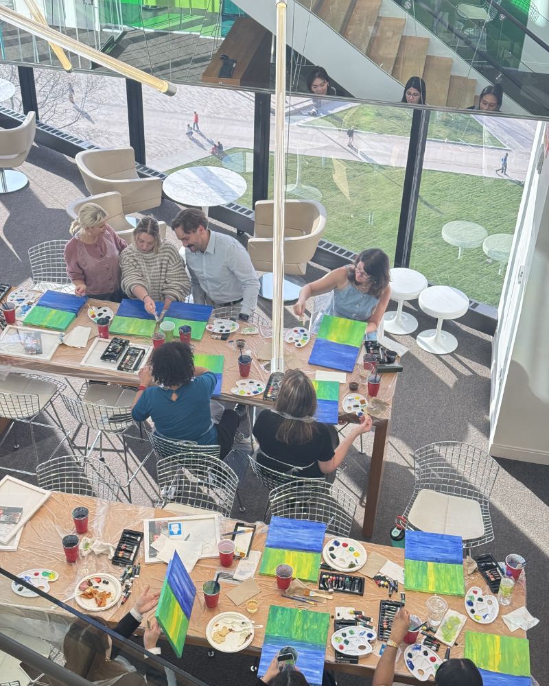 People seated around tables in a bright café or lounge, working and chatting by large windows.