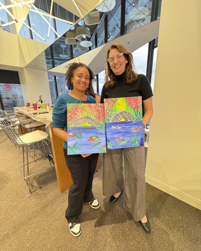 Two people in a bright lobby holding colorful painted canvases and smiling at the camera.