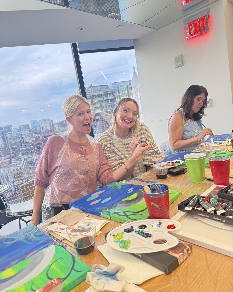 People painting at a table in a bright room with city views, colorful supplies spread out.