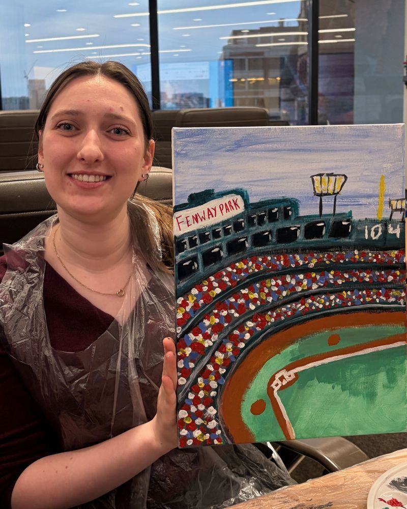 Smiling woman holding a colorful painting of a stadium scene indoors