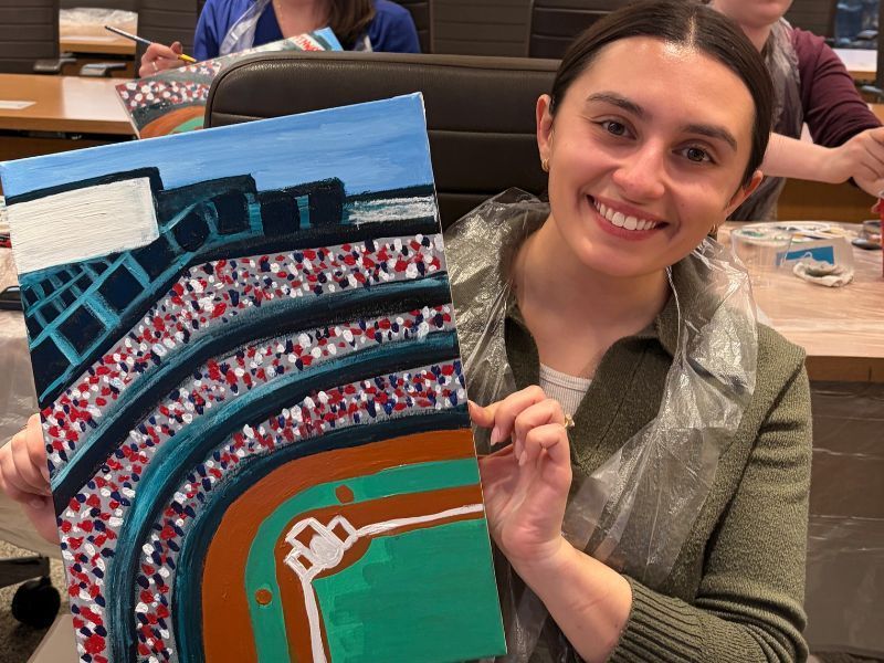 Smiling woman holding a painted stadium artwork with blue seating and a green field indoors