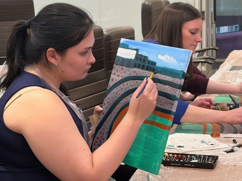 Two people painting on canvas at a table, with brushes and art supplies in a classroom or studio setting.