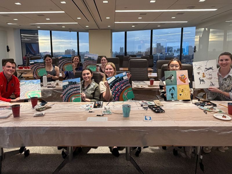 Group of people smiling at a long table with art supplies in a bright office lounge.