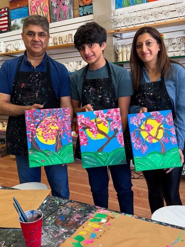 Family holding paintings of a tree, moon, and flowers. They wear aprons in a brightly lit art studio.