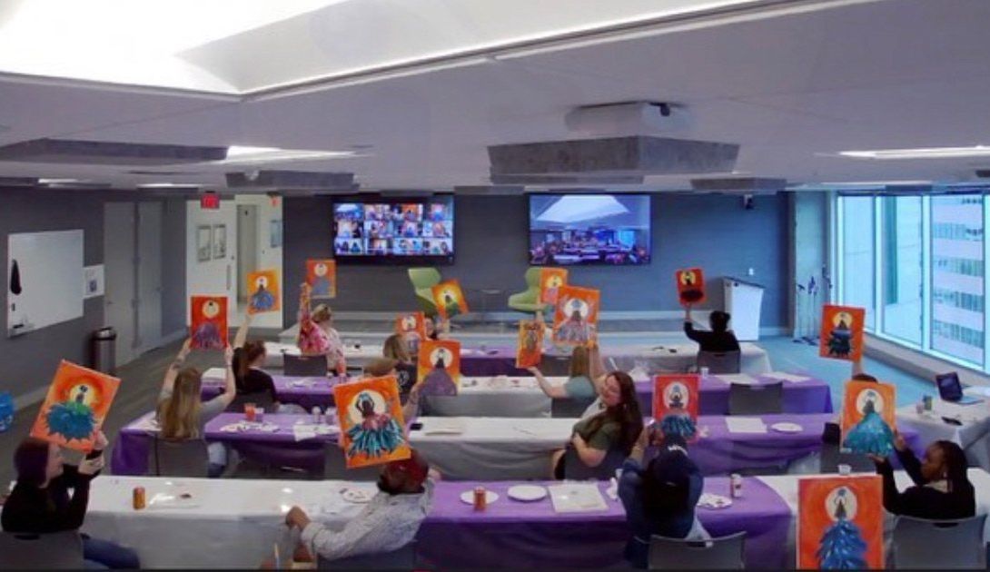 People in a room hold up paintings during a group art activity. Tables covered with purple cloths.
