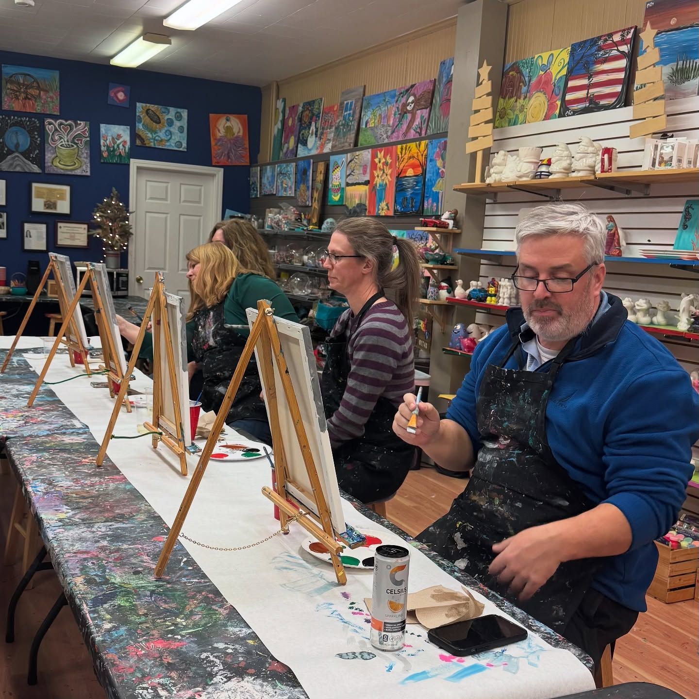 A group of people are sitting at a table painting on easels